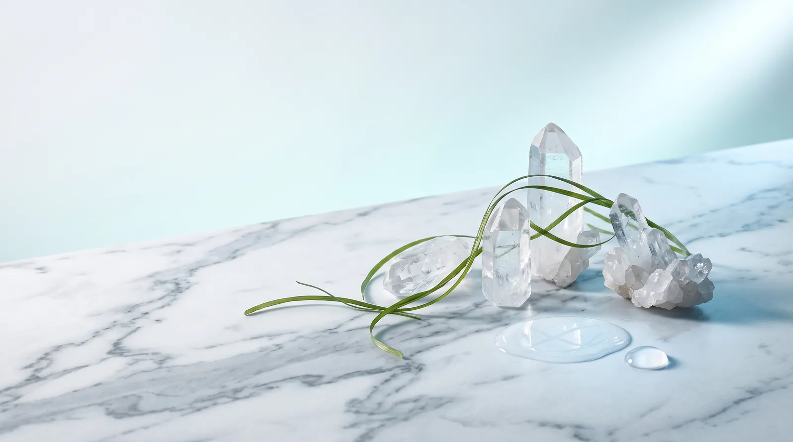 Glacier-blue hyaluronic still life — clear quartz points, a seagrass strand and a pool of water on white marble